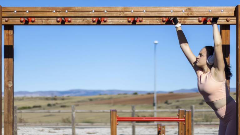 Porträt einer sportlichen Frau beim Klimmzug am Barren an einem sonnigen Tag mit Kopfhörern und Sportkleidung