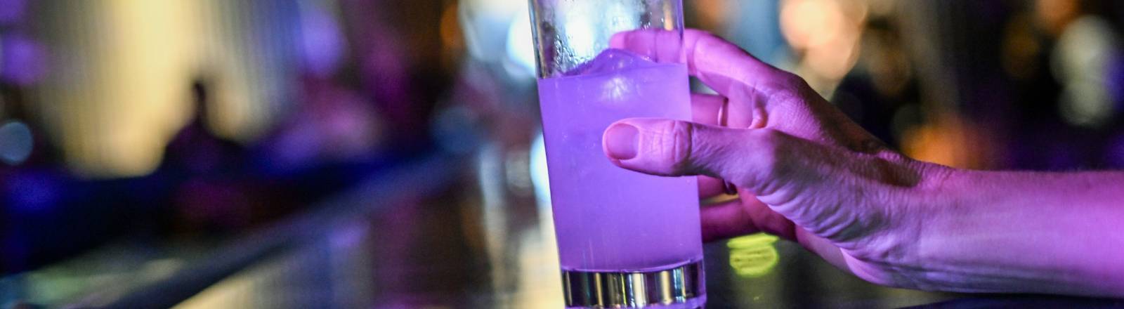 Eine Frau steht an einer Theke und greift nach einem Glas mit einem Cocktail, der violett leuchtet.