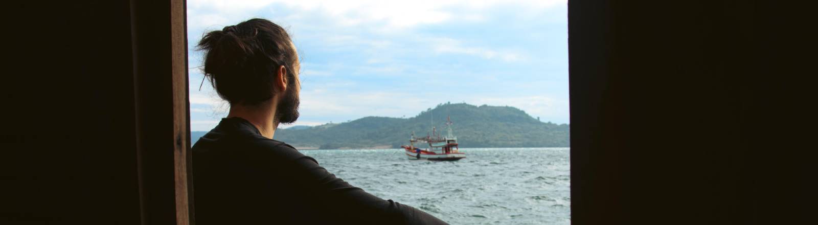 Ein Mann sitzt auf einem Steg und blickt auf das Meer und ein Boot mit Bergen im Hintergrund