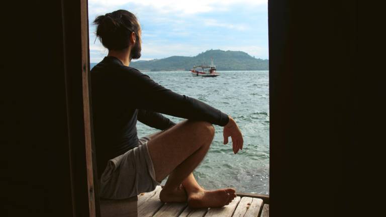 Ein Mann sitzt auf einem Steg und blickt auf das Meer und ein Boot mit Bergen im Hintergrund