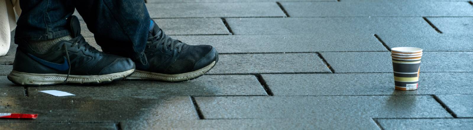 Schuhe eines Menschen, vor ihm steht ein Pappbecher