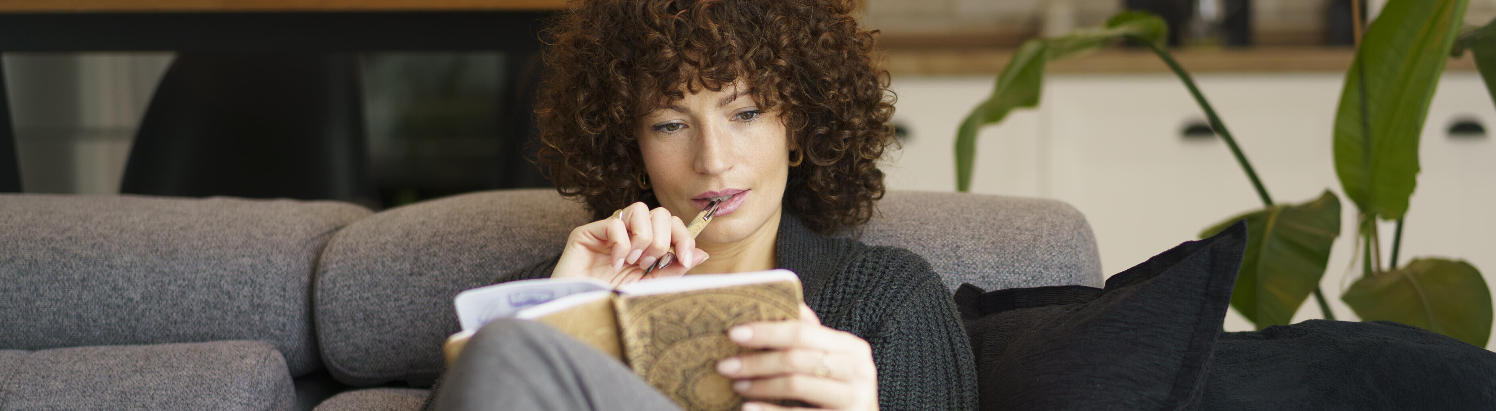 Frau sitzt mit angezogenen Knien auf Sofa mit Notizbuch, Stift im Mund und nachdenklichem Blick