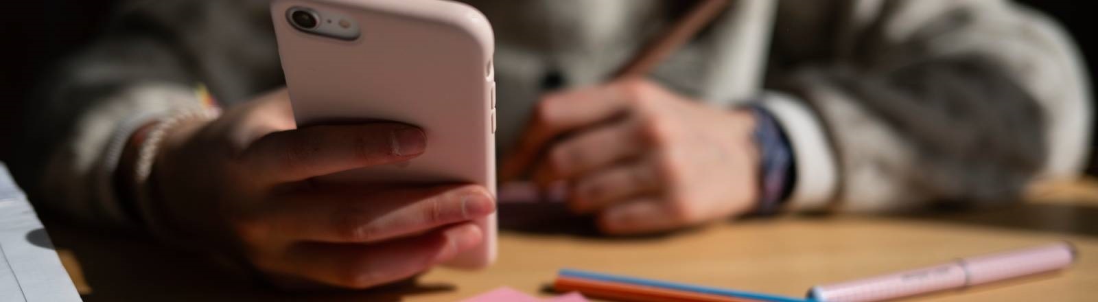 Ein Schulkind sitzt am Schreibtisch, bearbeitet Aufgaben mit einem Stift und hält dabei ein Smartphone in der Hand.