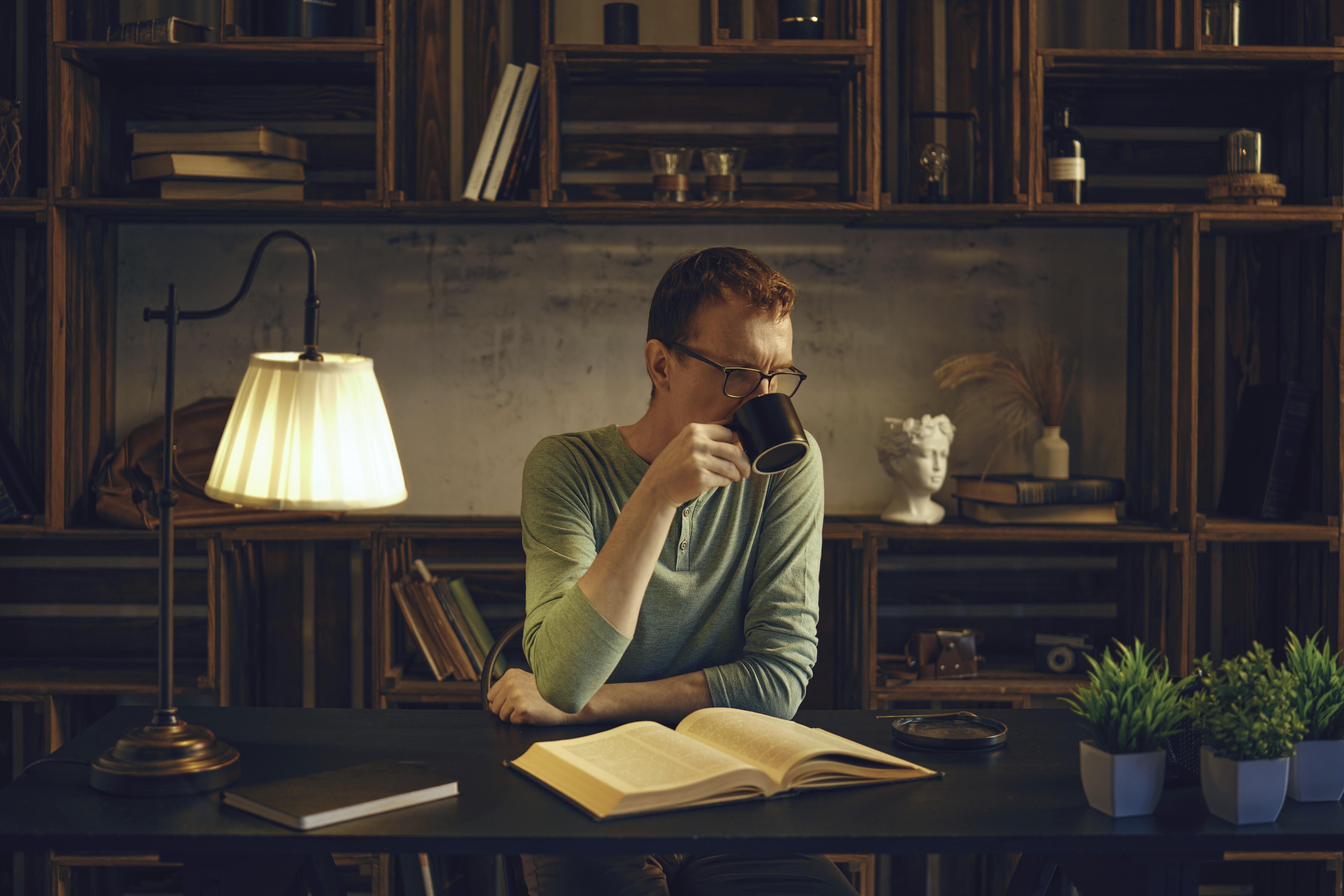 Ein Mann mittleren Alters sitzt an einem Schreibtisch in einem gemütlichen Zimmer und schlürft einen Kräutertee aus einer Tasse. Das sanfte Licht einer Lampe beleuchtet das Buch, das er vor dem Schlafengehen liest