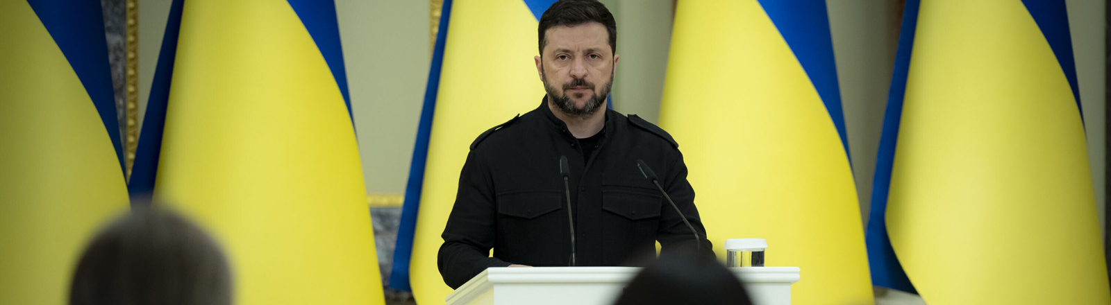Der ukrainische Präsident Volodymyr Selenskyj steht am Mikrofon, hinter stehen ukrainische Flaggen in einer langen Reihe