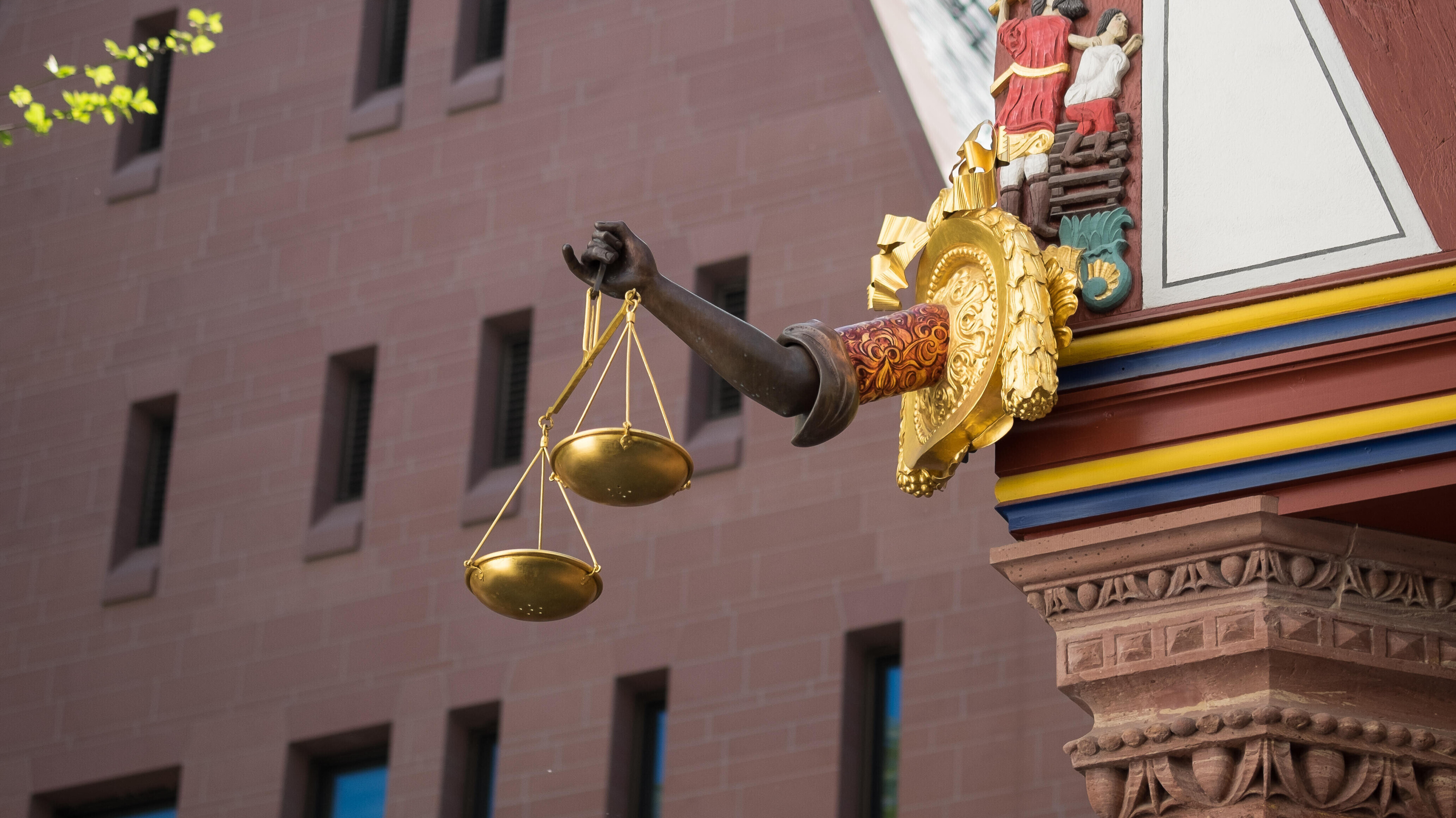 Ein ausgestreckter Arm aus Holz hält eine goldene Waage an einem rekonstruierten Fachwerkhaus