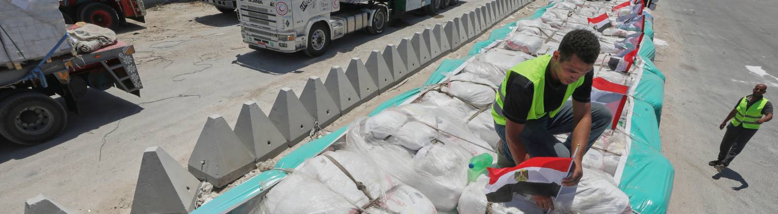 Mann mit gelber Warnweste befestigt ägyptische Flaggen auf Hilfslieferungen, die auf einem LKW liegen. Mehrere solcher LKWs stehen an der Grenze Gaza-Ägypten (06.08.2025)