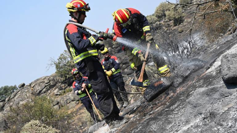 Deutsche Feuerwehrleute spritzen in der Region Extremadura Wasser auf einen bereits abgebrannten Bereich und suchen nach möglichen Glutnestern. Sie tragen Schutzkleidung und Helme. Die Pflanzen sind verkohlt und Felsen schwarz.
