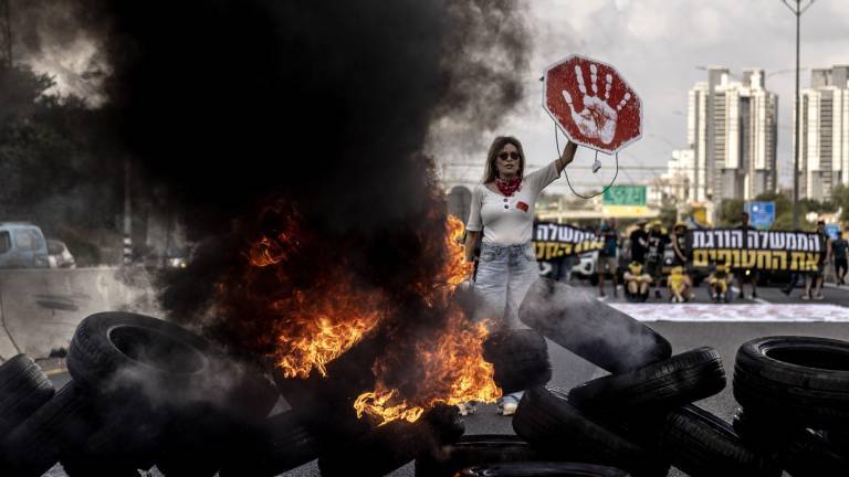 Demonstrantin in Tel Aviv (Israel) hält ein rotes Schild mit weißem Handabdruck in die Höhe; vor ihr liegt eine Barrikade aus brennenden Reifen, neben ihr steigt eine schwarze Rauchwolke in die Höhe (26.08.2025)