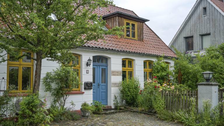Ein Wohnhaus in einem ländlichen Stil von vor 100 Jahren. Vor dem Haus ist ein kleiner Garten mit Blumen zu sehen.