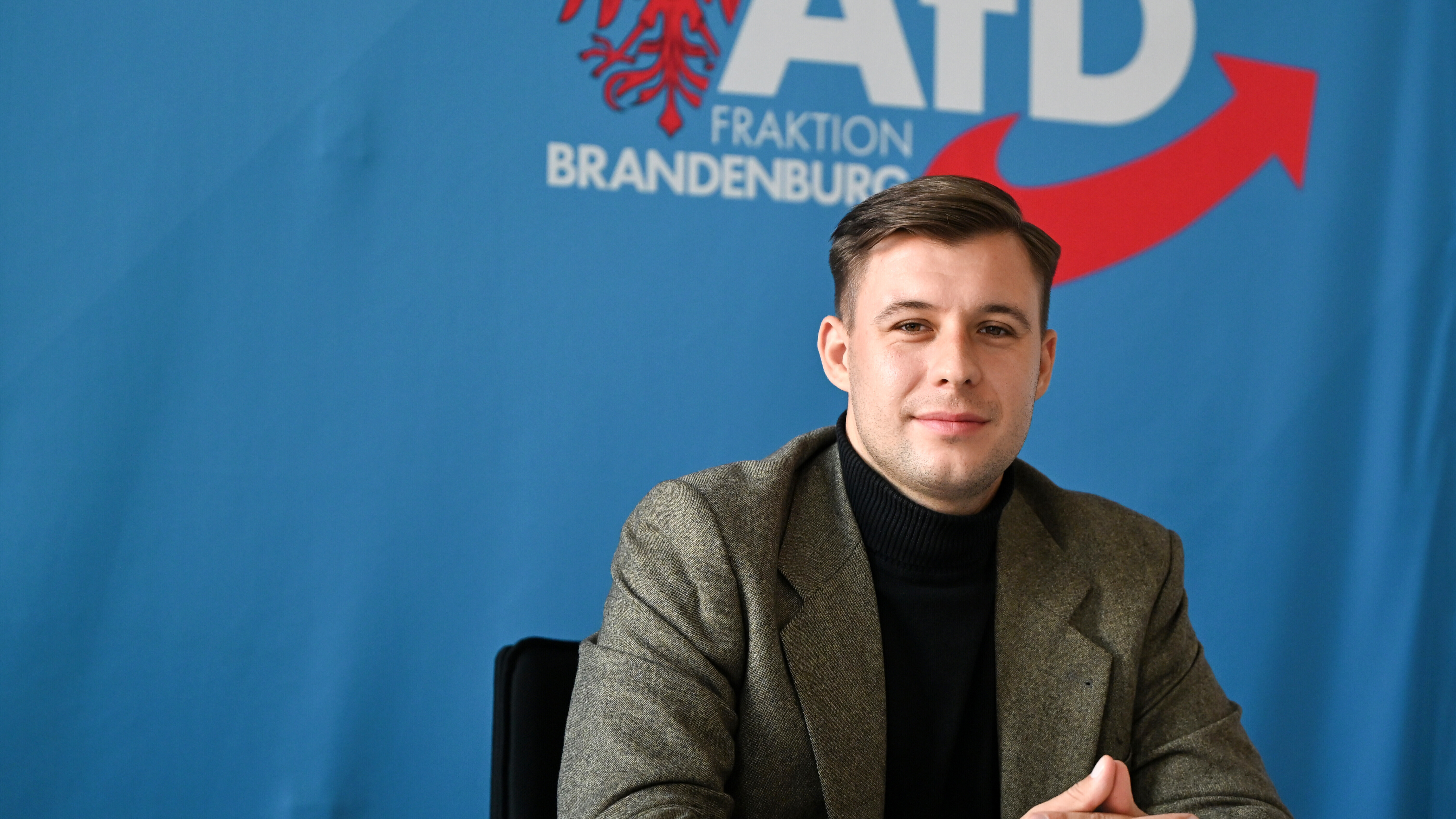 Jean-Pascal Hohm, AfD-Landtagsabgeordneter Brandenburg und Anwärter auf den Vorsitz der neuen AfD-Jugend, sitzt vor einem blauen Banner mit dem Logo der AfD-Brandenburg.