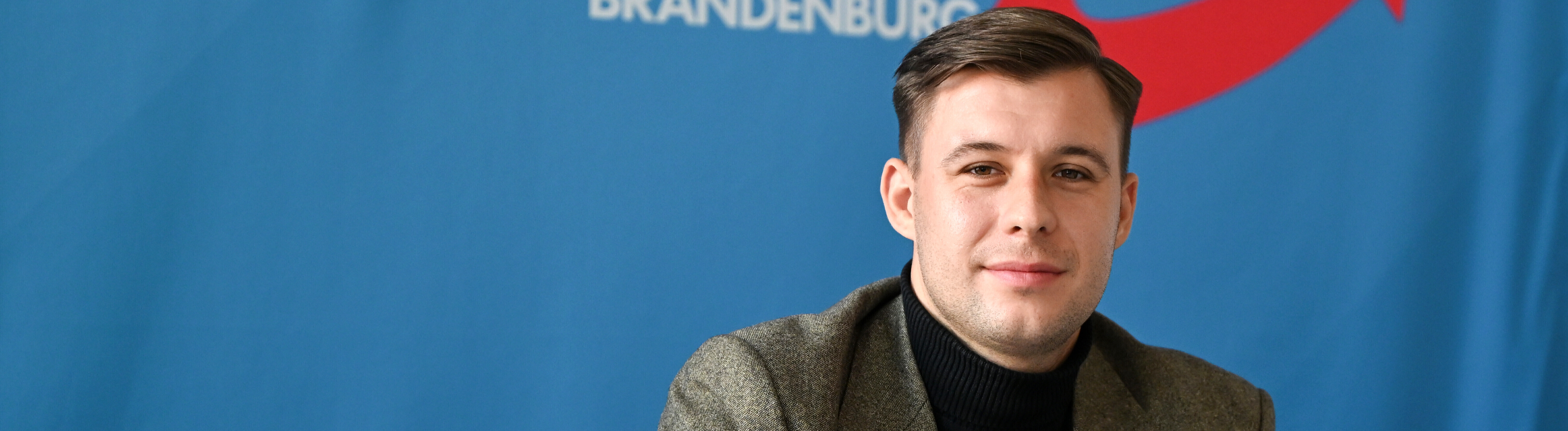 Jean-Pascal Hohm, AfD-Landtagsabgeordneter Brandenburg und Anwärter auf den Vorsitz der neuen AfD-Jugend, sitzt vor einem blauen Banner mit dem Logo der AfD-Brandenburg.