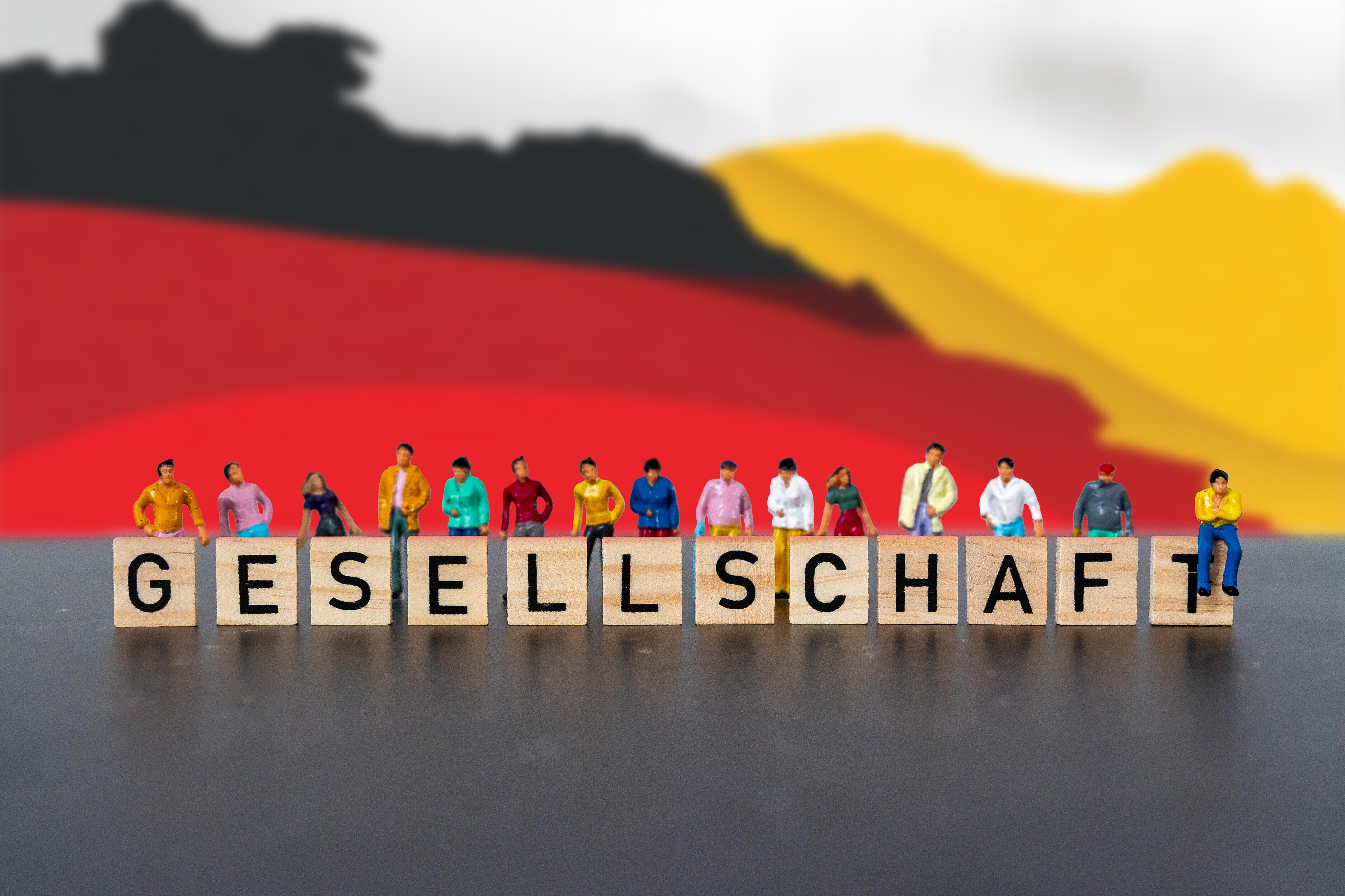 Fotomontage, Menschen stehen hinter Holzbuchstaben mit dem Wort „Gesellschaft“ vor einer Deutschland-Flagge /  Sinnbild für nationale Identität, Vielfalt, Zusammenhalt und die Bedeutung gemeinsamer Werte in Deutschland.