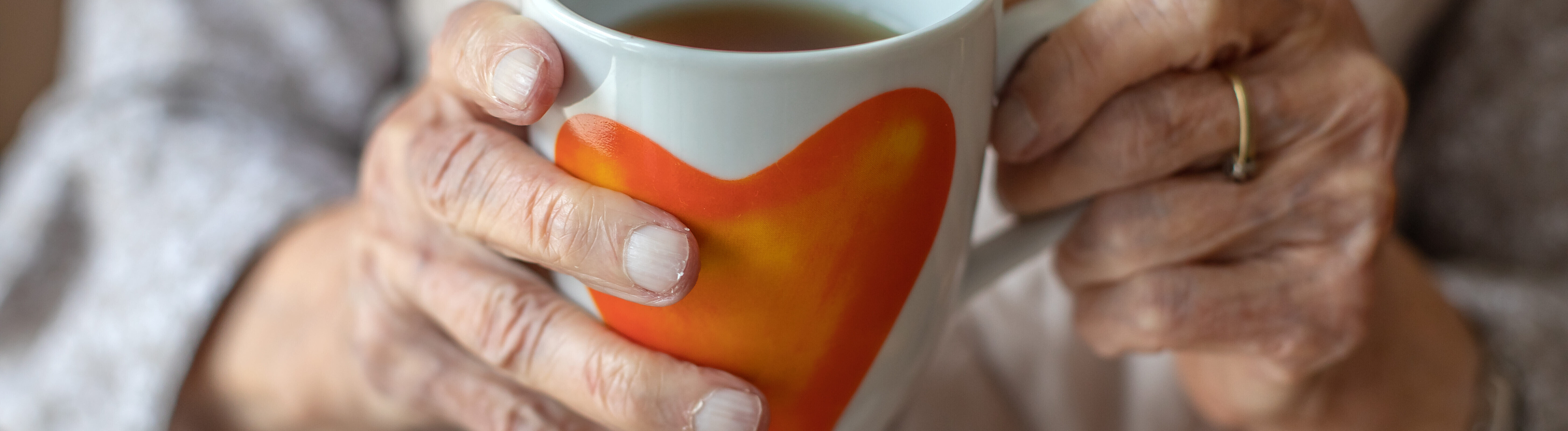 Die Hände einer älteren Frau halten eine Teetasse mit Herzmotiv.