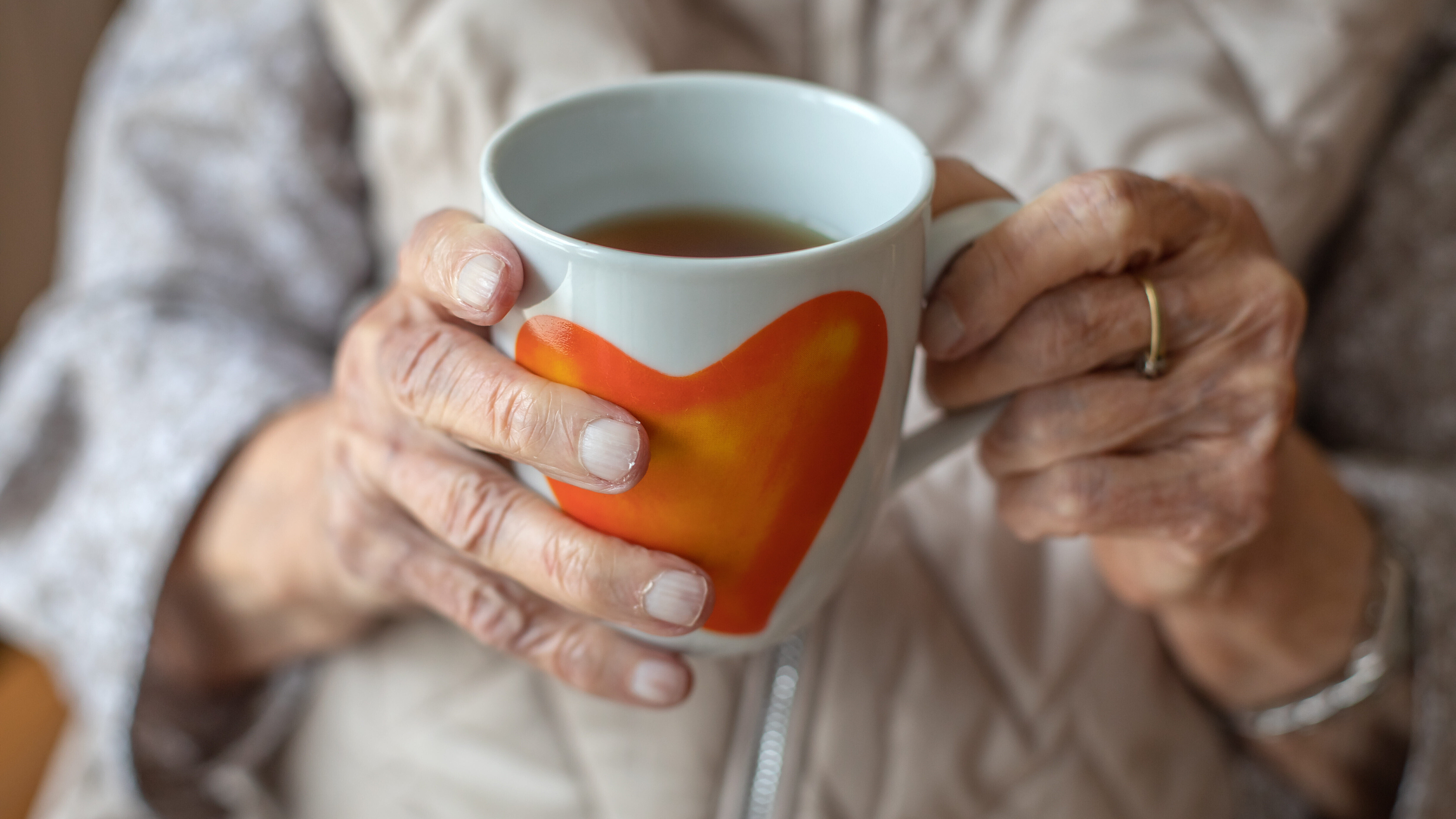 Die Hände einer älteren Frau halten eine Teetasse mit Herzmotiv.
