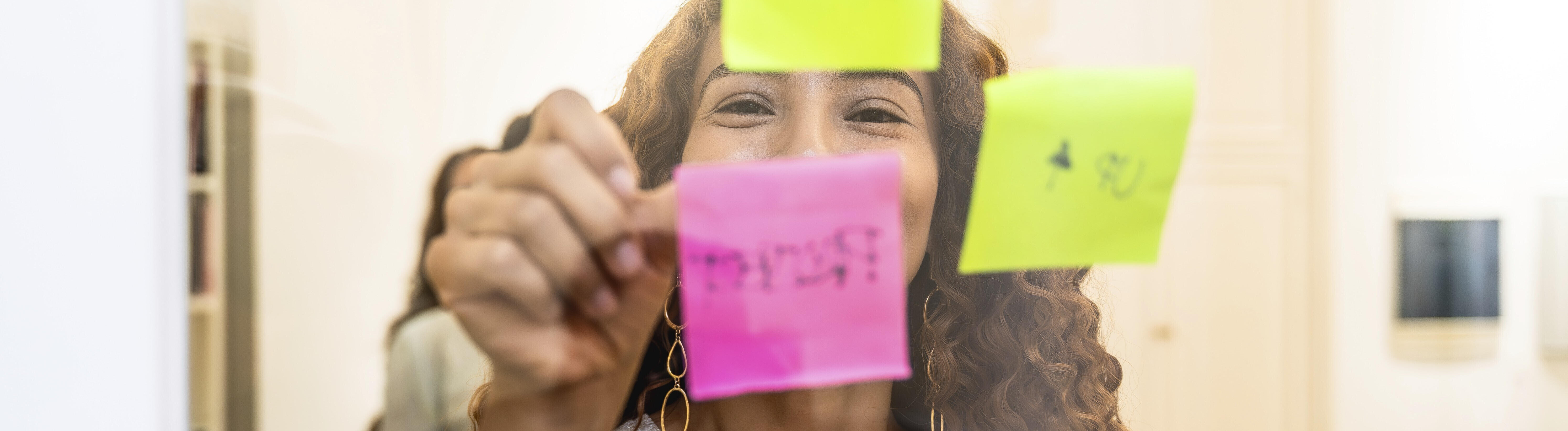 Eine Frau schreibt lächelnd Ideen und Aufgaben auf bunte Haftnotizen an einer klaren Glaswand.