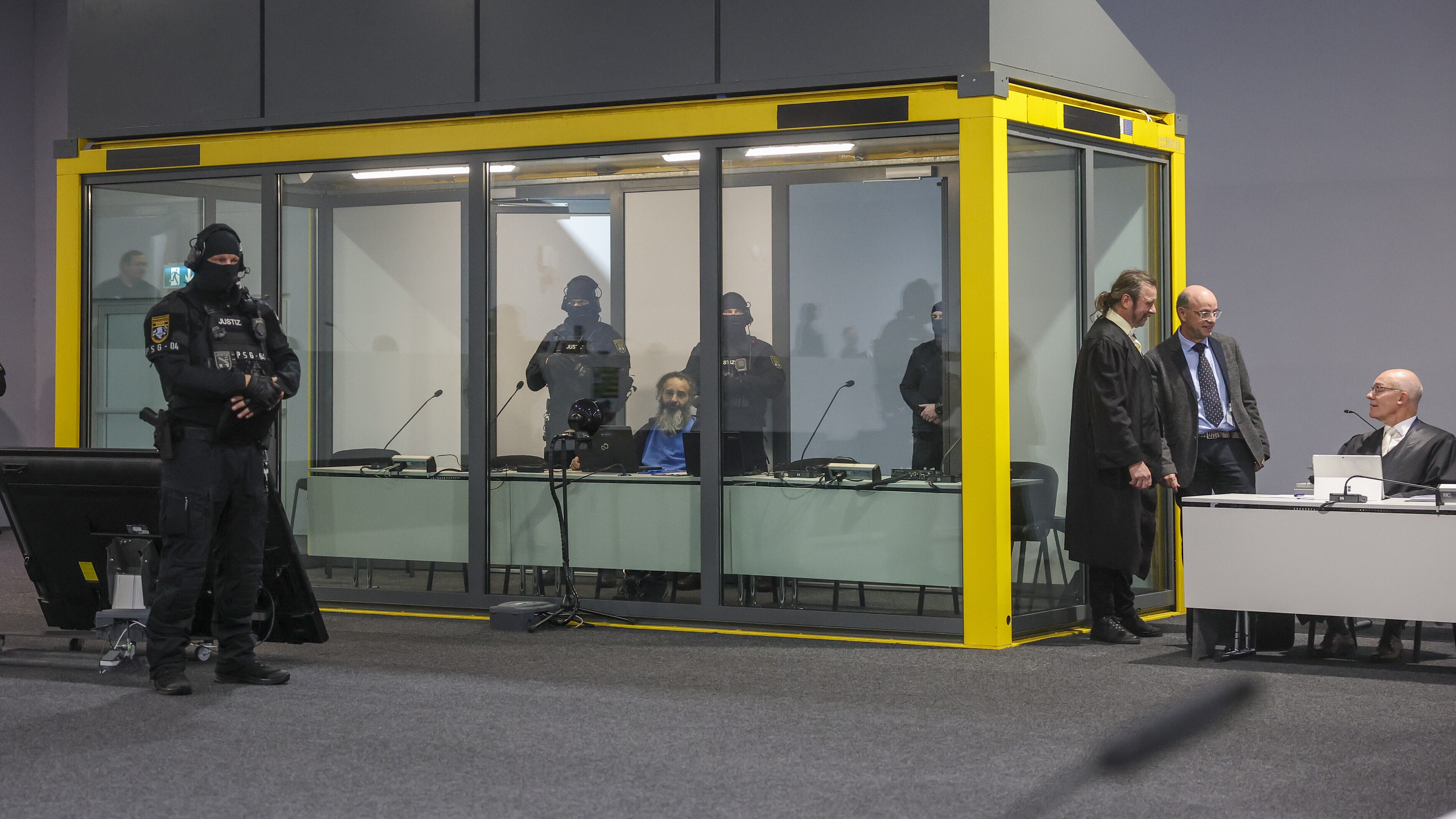 Hinter einem Glaskasten sitzt der Angeklagte, um ihn Polizisten