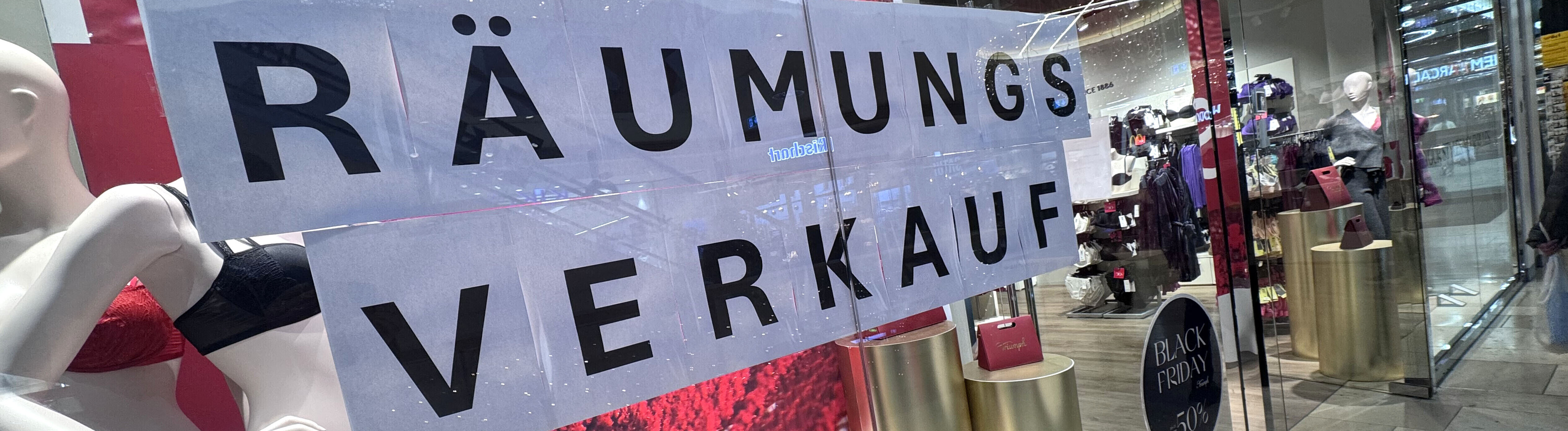 In einem Schaufenster hängt ein Banner mit der Aufschrift "Räumungsverkauf" dahinter befinden sich Schaufensterpuppen in Unterwäsche und das Logo der Firma Triumph  ist zu lesen.