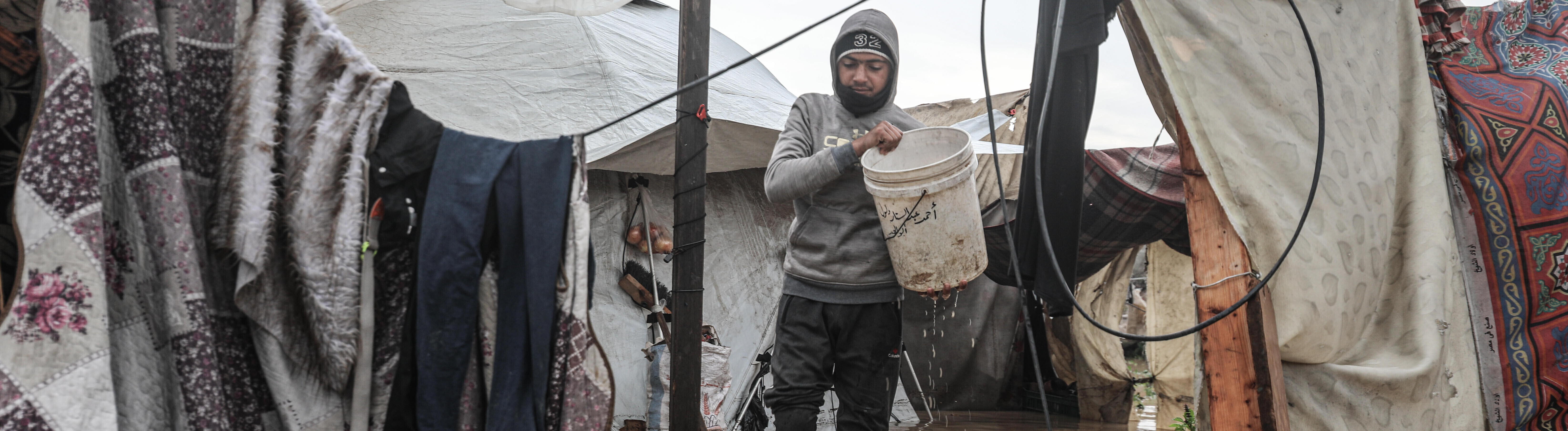 Mann, der einen Eimer hält, geht auf einem überfluteten Platz an Zelten vorbei (12.12.2025, Deir Al-Balah in Gaza)