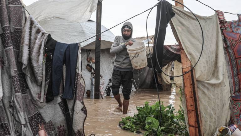 Mann, der einen Eimer hält, geht auf einem überfluteten Platz an Zelten vorbei (12.12.2025, Deir Al-Balah in Gaza)