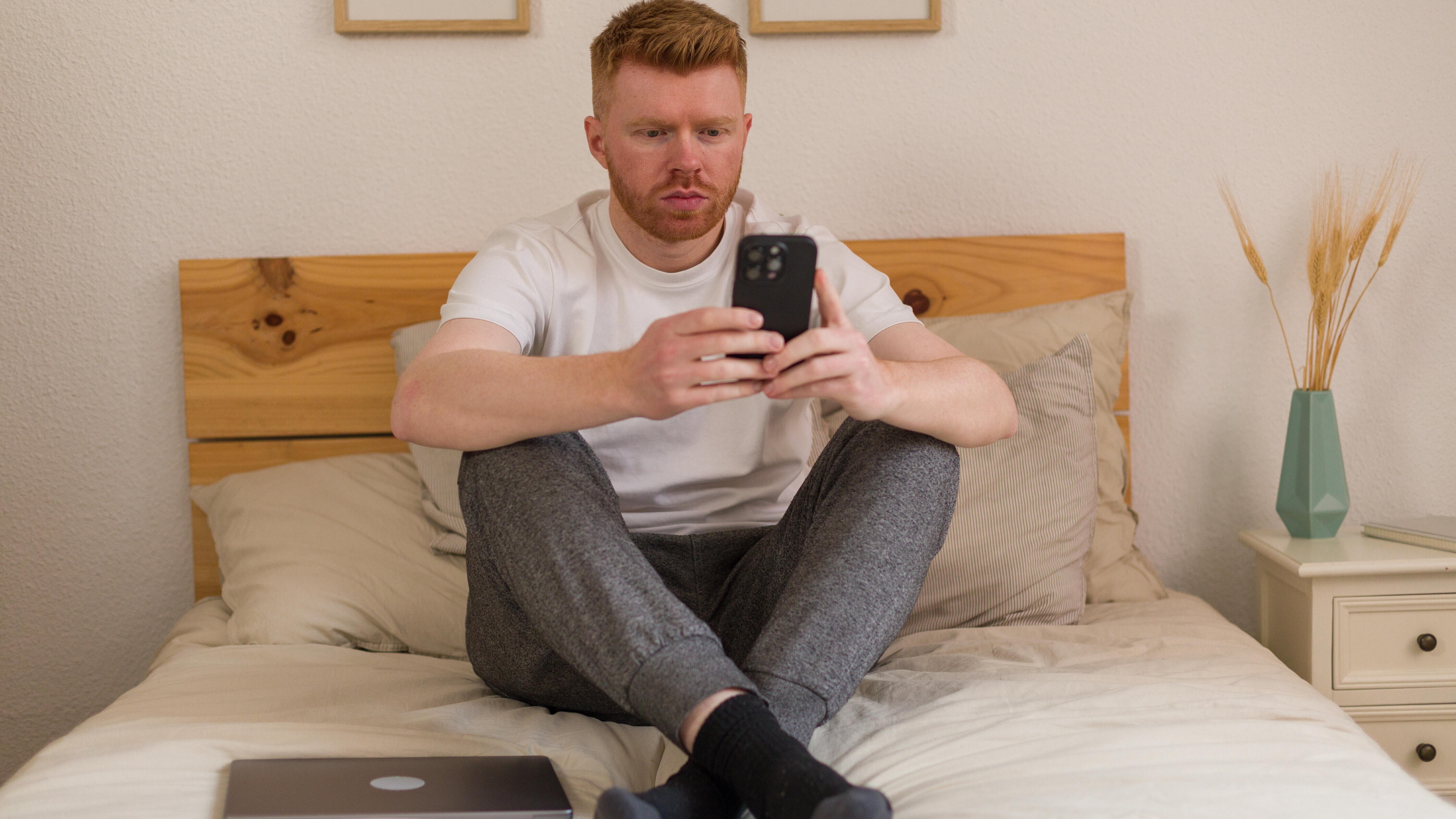 Mann sitzt auf Bett und schaut konzentriert aufs Handy, neben ihm liegt ein Laptop