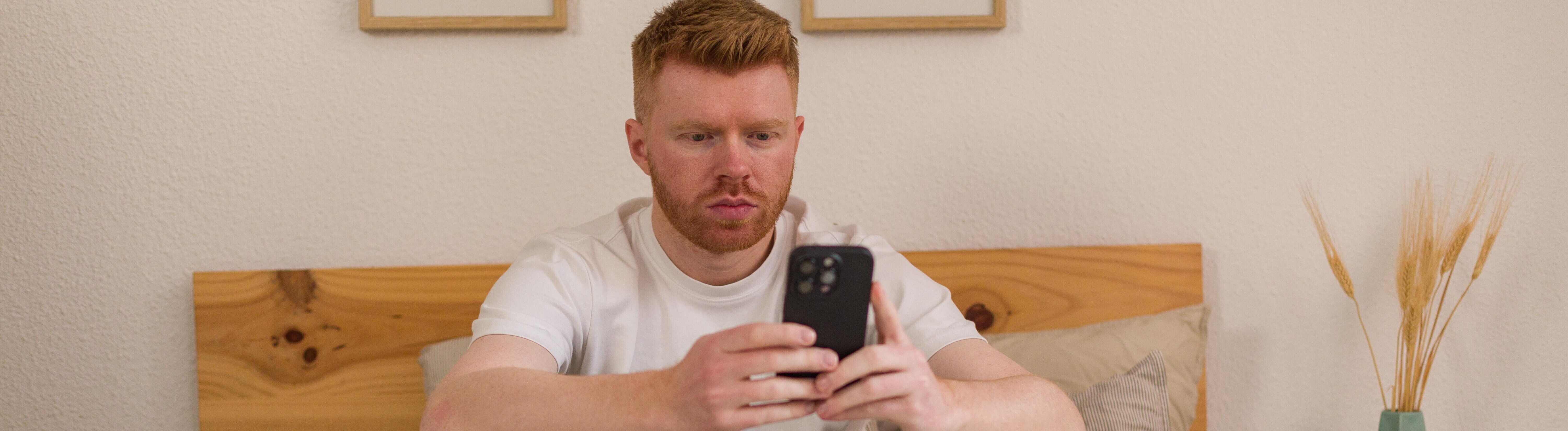 Mann sitzt auf Bett und schaut konzentriert aufs Handy, neben ihm liegt ein Laptop