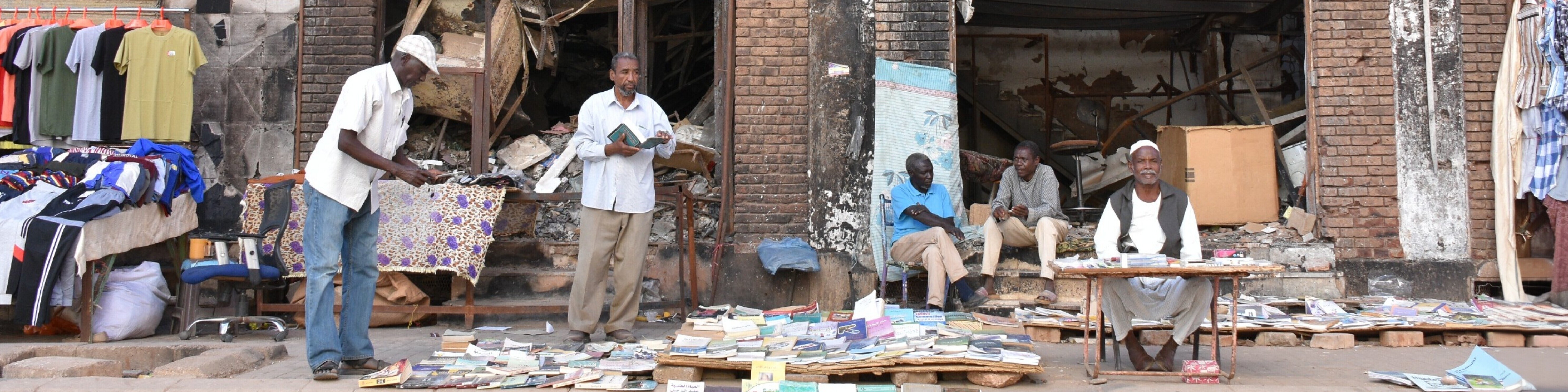 In der sudanesischen Hauptstadt Khartum herrscht auf der Straße wieder Alltag: Händler verkaufen Kleidung und Bücher, hinter ihnen steht ein Gebäude mit schwer beschädigter Fassade.