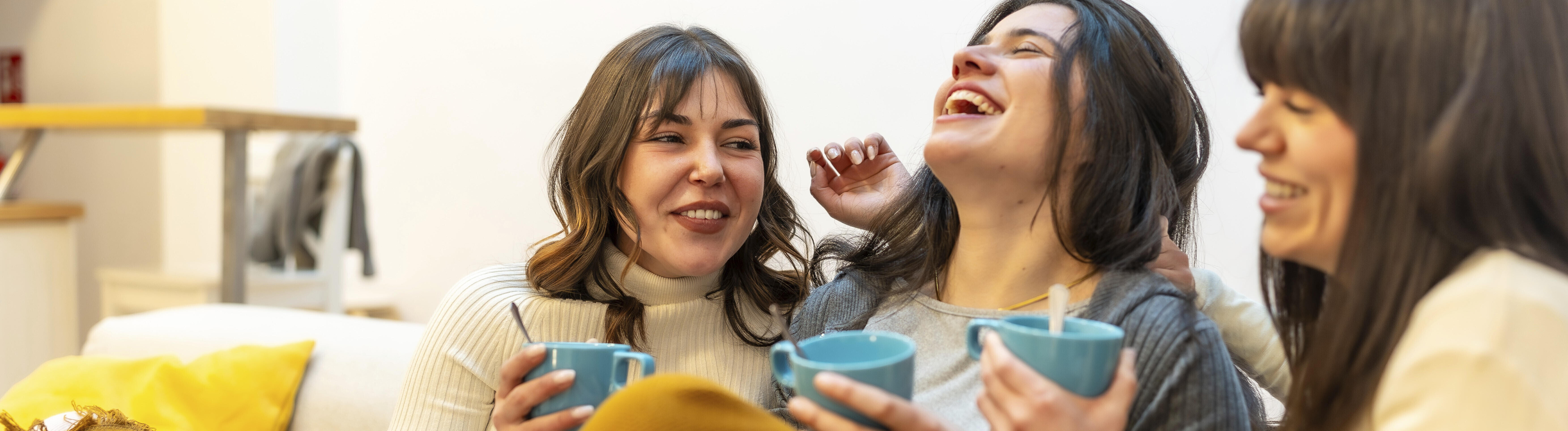 Glückliche Freundinnen, die zu Hause einen gemütlichen Moment zu dritt genießen, lachen und heiße Getränke trinken, während sie auf der Couch unter einer warmen Decke sitzen