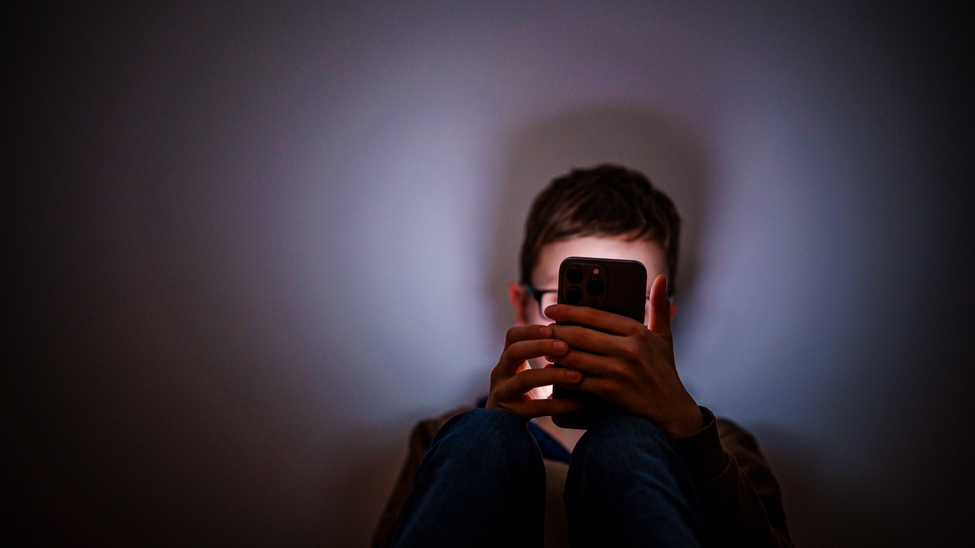 Ein junger Mann sitzt im Dunkeln und schaut auf sein Smartphone, das Licht vom Display leuchtet auf sein gesicht, das jedoch vom Smartphone verdeckt ist.