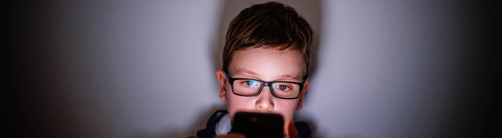 Ein Junge sitzt in einem dunklen Raum und wird von einem Smartphone beleuchtet. Symbolfoto. Gestellte Aufnahme zum Thema Kinder nutzen Social Media.