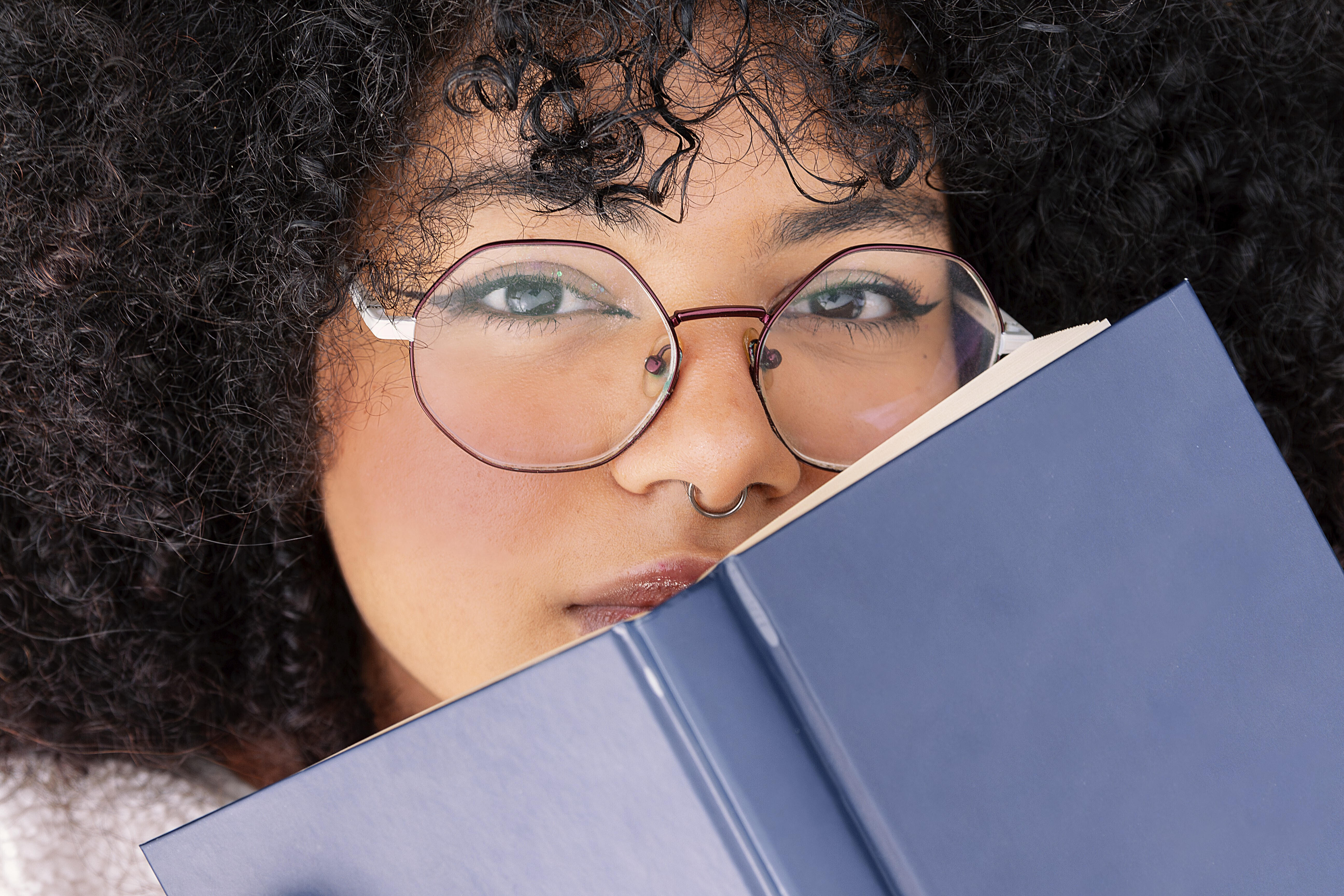 Eine Frau mit lockigem Haar schaut über ein blaues Buch und trägt eine große runde Brille. Ihr Gesichtsausdruck ist konzentriert und unterstreicht ein Gefühl von Neugier und Kontemplation