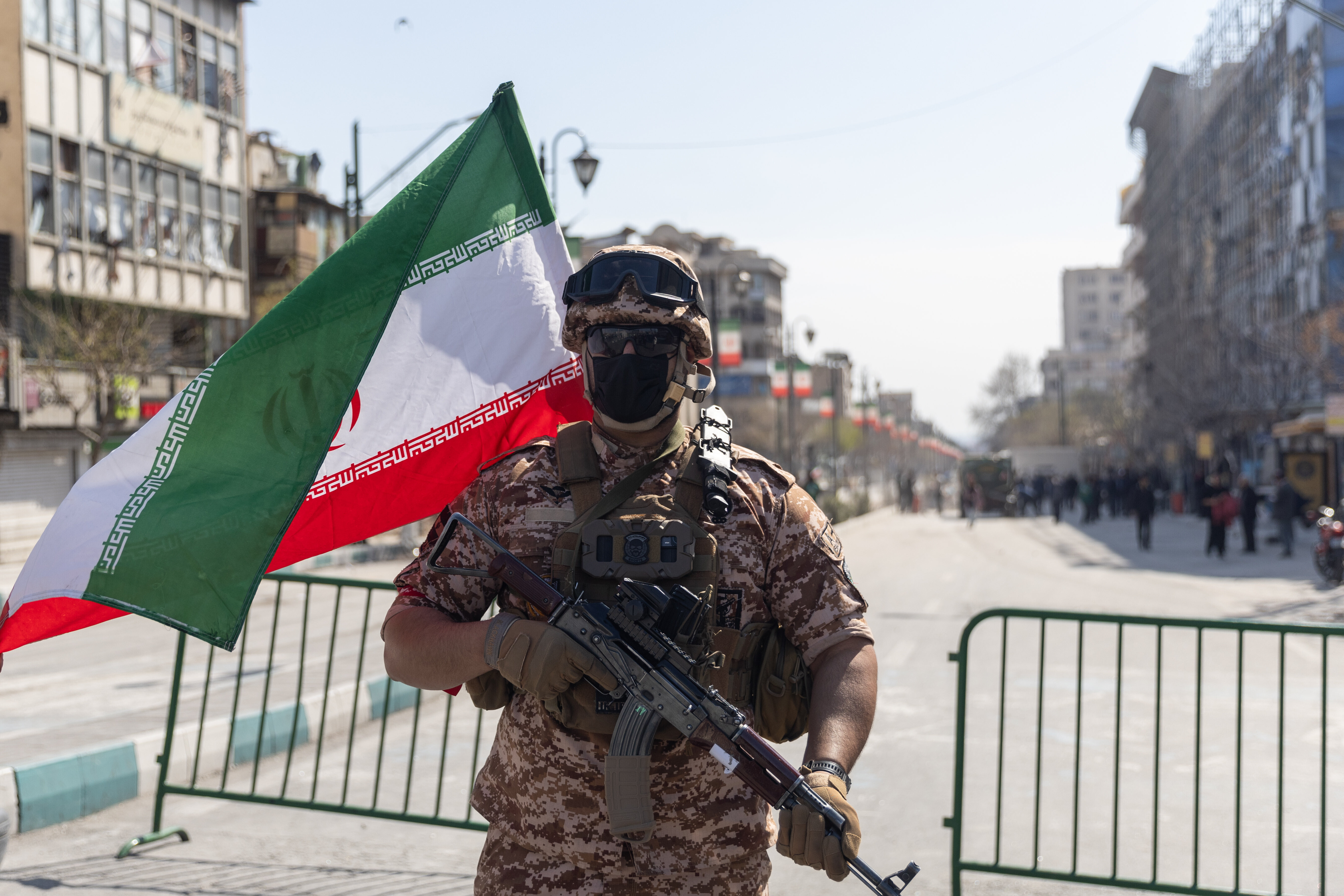 Ein Soldat steht in Iran auf einer Straße und hält ein Maschinengewehr. Hinter ihm steht ein Gitter, eine Iran-Flagge weht im Wind.