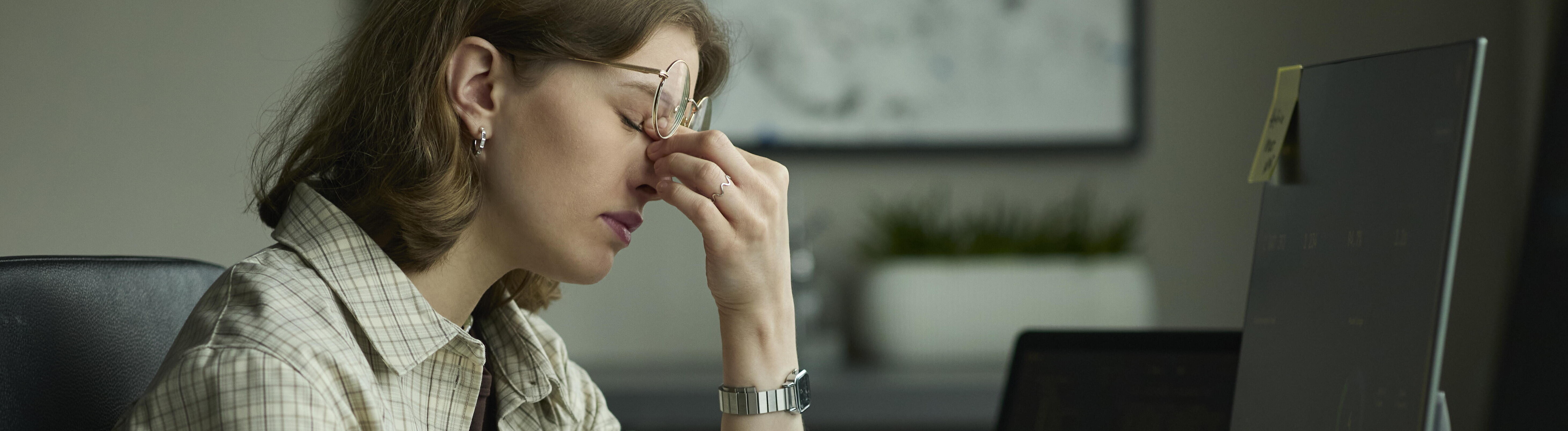 Eine junge Frau sitzt an ihrem Arbeitsplatz vor einem Computer. Sie wirkt müde und drückt mit zwei Fingern auf ihre Nasenwurzel. Dabei rutscht ihre Brille hoch.