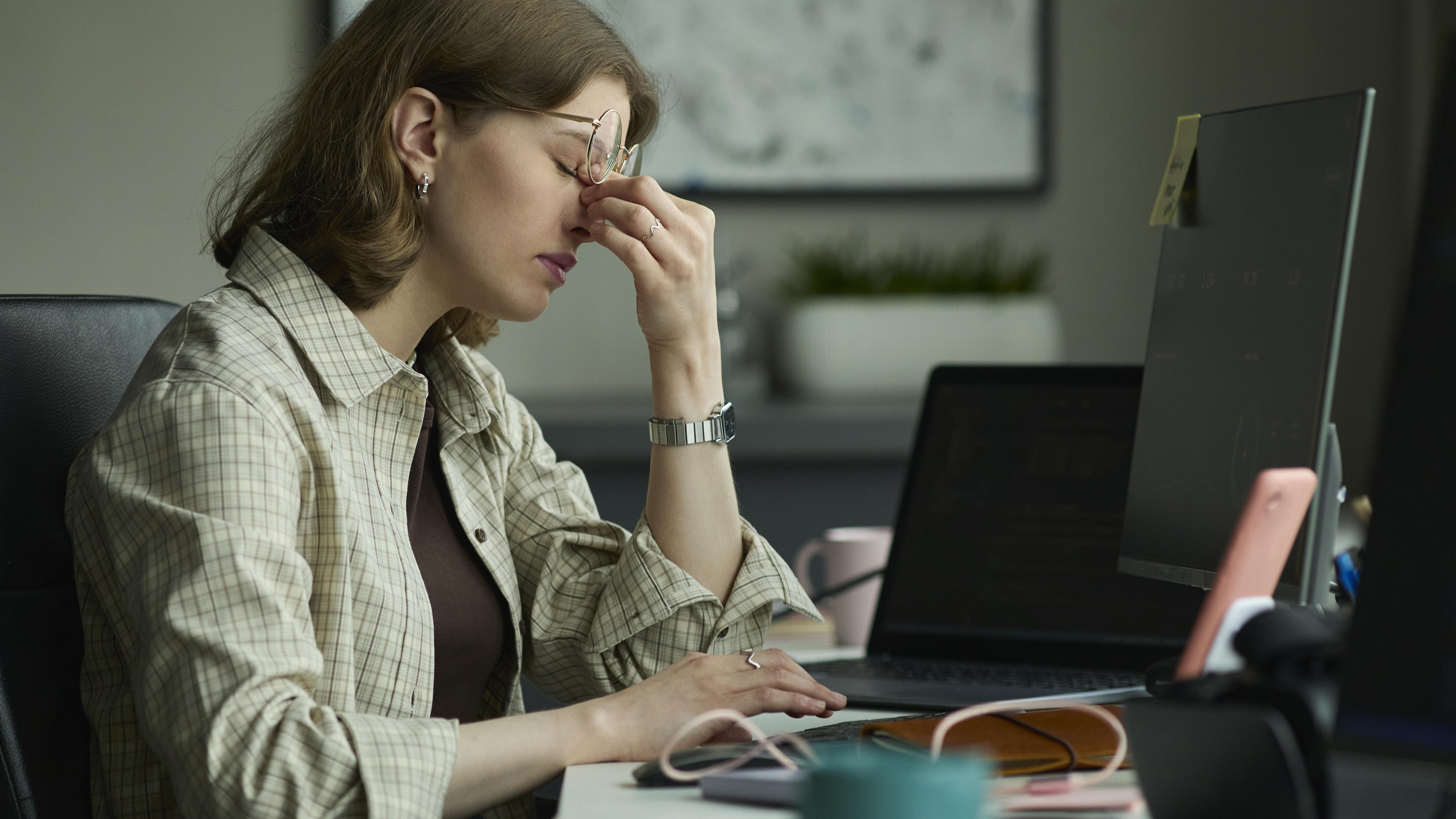 Eine junge Frau sitzt an ihrem Arbeitsplatz vor einem Computer. Sie wirkt müde und drückt mit zwei Fingern auf ihre Nasenwurzel. Dabei rutscht ihre Brille hoch.