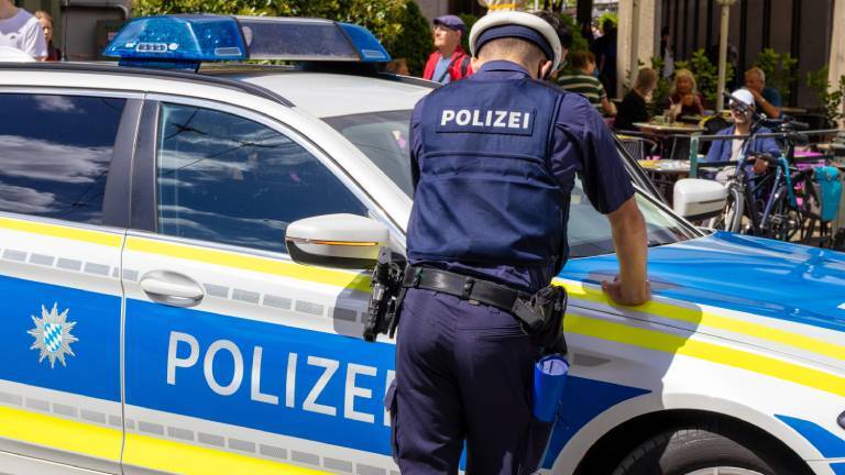 Ein Polizist lehnt an seinem Polizeiwagen in Augsburg. Er trägt Uniform. Im Hintergrund ist eine Fußgängerzone zu sehen.