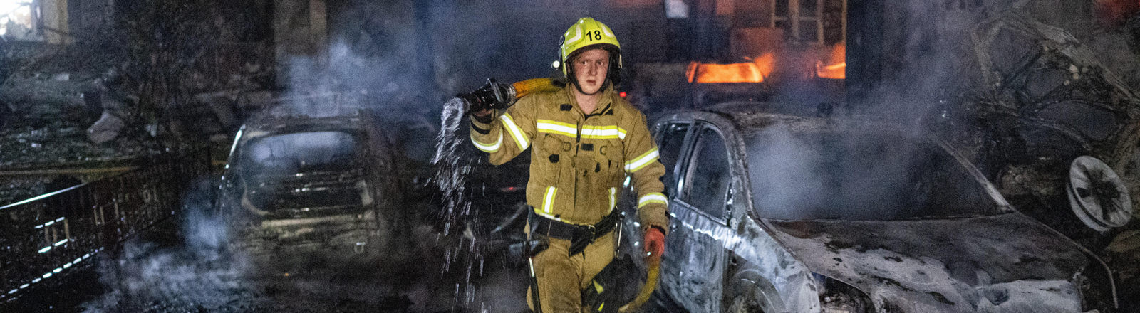 Nach einem russischen Raketenangriff in Kiew versucht eine Feuerwehrmann Brandherde zu löschen. Neben ihm stehen ausgebrannte Autos. Im Hintergrund lodern Feuer in einem Wohnhaus.