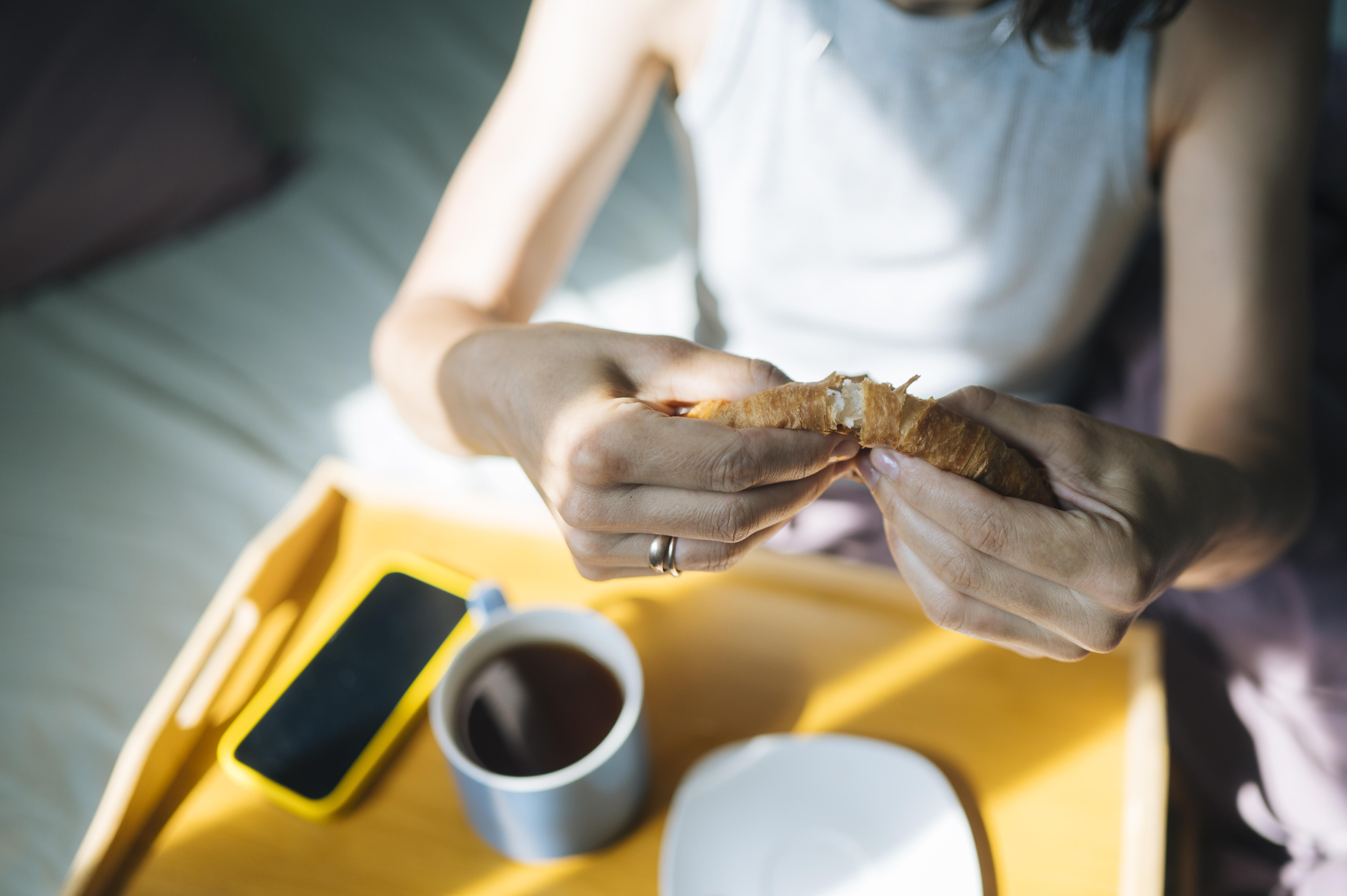 Eine Frau teil ihr Croissant während ihres Frühstücks im Bett. Das Handy liegt parat und eine Tasser Kaffee steht auch herum