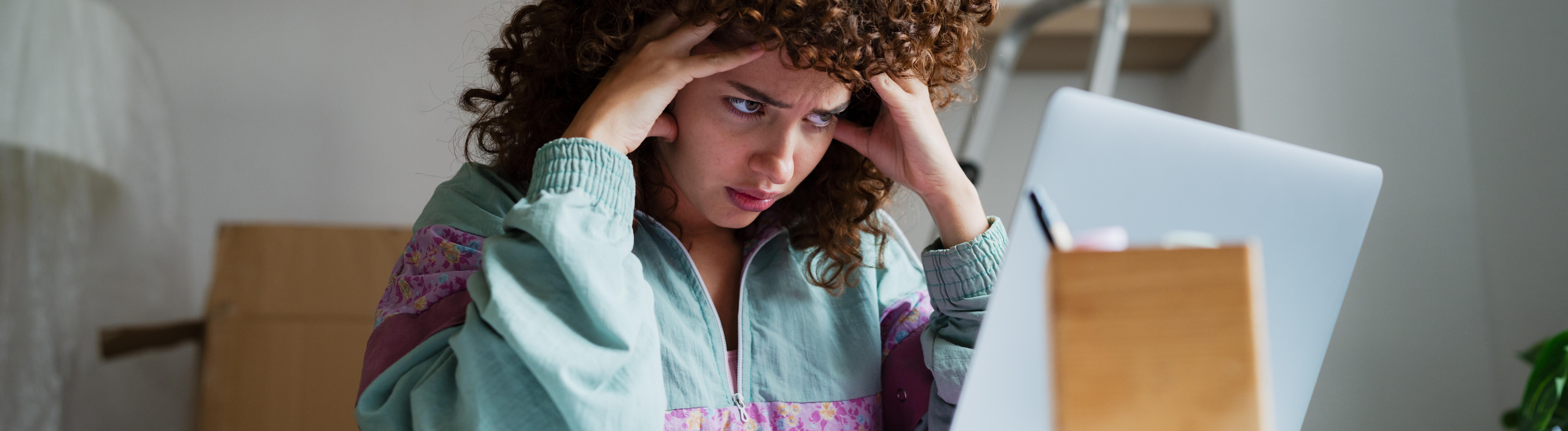 Eine Frau sitzt inmitten von Umzugskartons an einem Schreibtisch. Sie schaut auf ihren Laptop, hat ihren Kopf auf ihre Hände gestützt und wirkt gestresst.