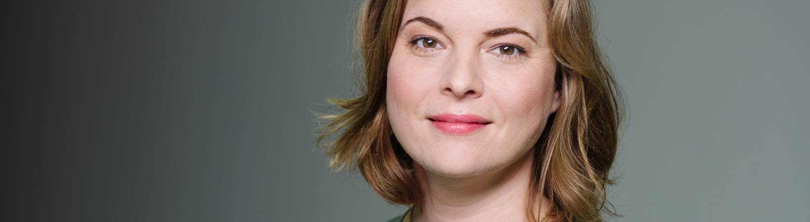 Portrait von Katja Berlin im olivegrünen Oberteil vor grauer Wand.