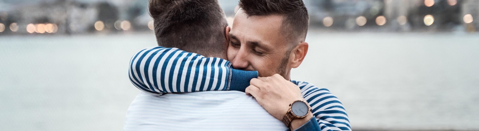 Ein Liebespaar umarmt sich. Im Hintergrund ein Hafenbecken und unscharf eine Stadt.