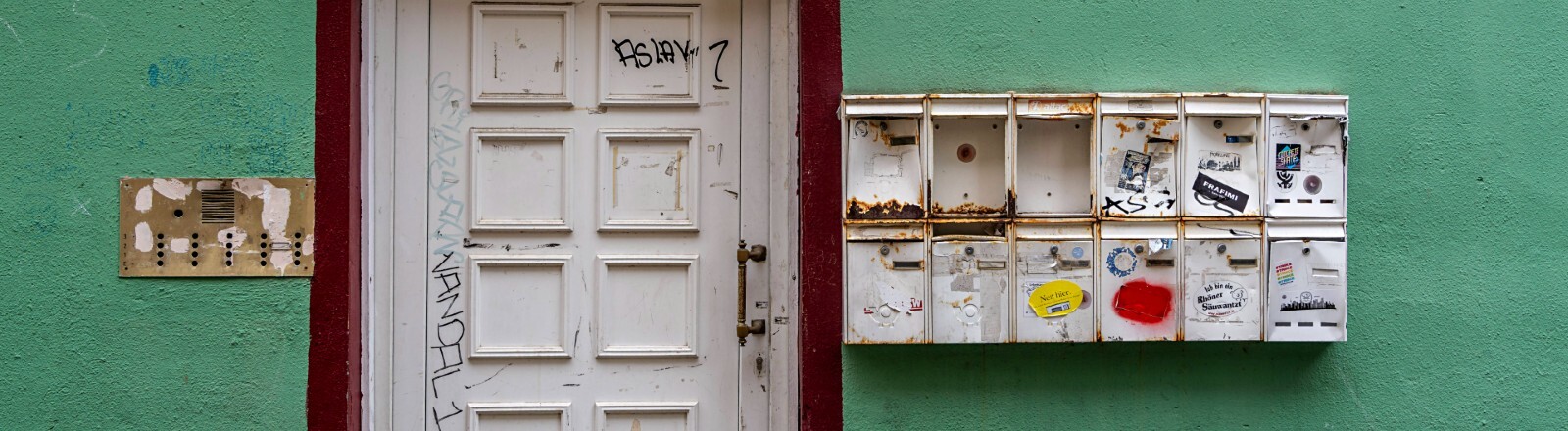 Eine alte weiße Haustüre eines grünen Hauses. Rechts daneben befinden sich kaputte, rostige Briefkästen, links ein lädiertes goldenes Klingelschild. Das Haus steht im Stadtteil Alt-Sachsenhausen in Frankfurt am Main.