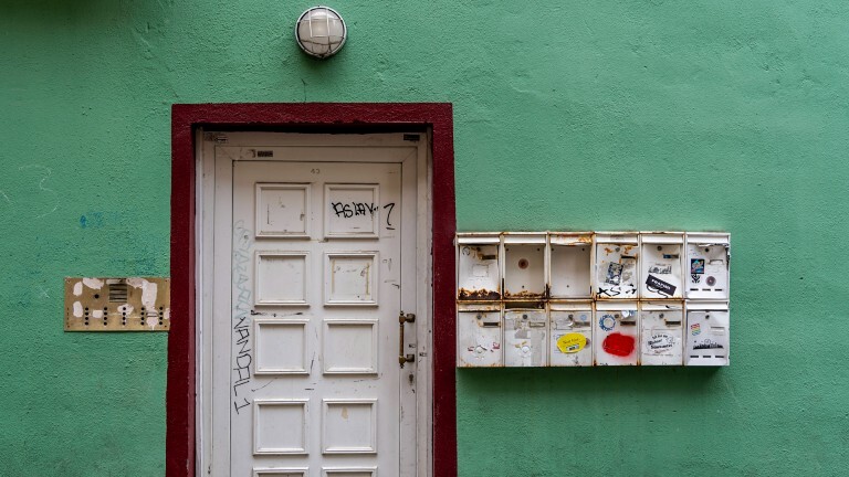 Eine alte weiße Haustüre eines grünen Hauses. Rechts daneben befinden sich kaputte, rostige Briefkästen, links ein lädiertes goldenes Klingelschild. Das Haus steht im Stadtteil Alt-Sachsenhausen in Frankfurt am Main.