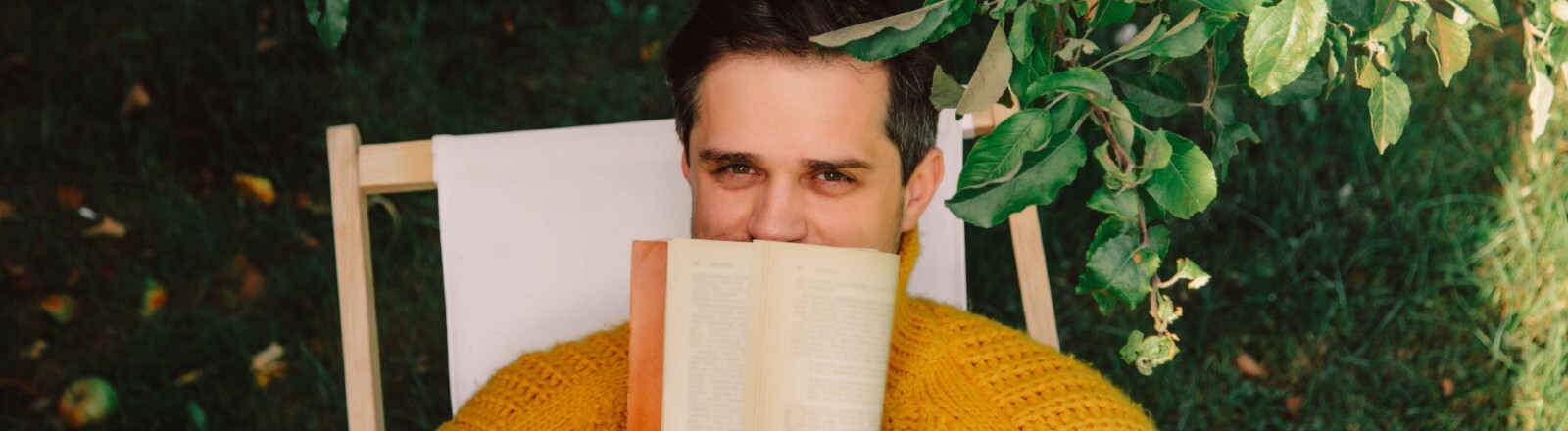 Ein Mann sitzt draußen in einem Liegestuhl unter einem Apfelbaum. Er hält sich ein Buch vor den Mund. Die Hälfte seines Gesichts ist dadruch verdeckt.