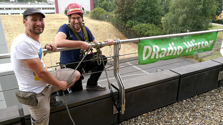 Industriekletterer Johannes Müller (l.) erklärt DRadio-Wissen-Moderator Sebastian Sonntag, was er zu tun hat