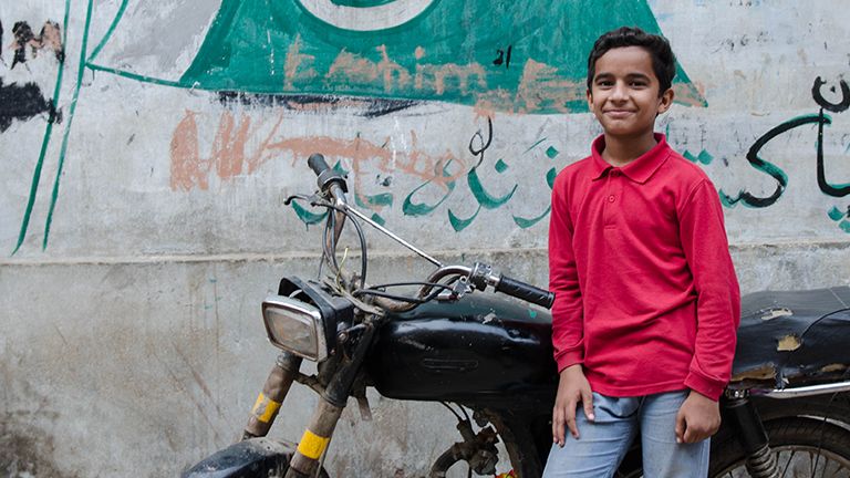 Ein Junge in einer Pakistanischen Straße posiert vor einem Motorrad.
