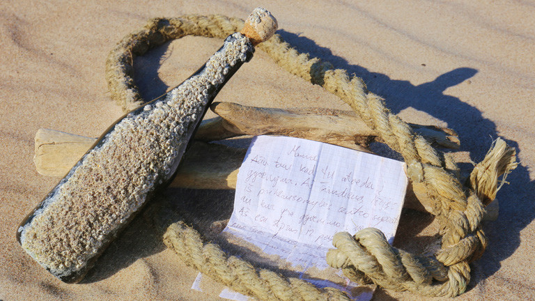 An Strand im Sand liegt ein Seil, darauf liegt ein Zettel und eine Flasche, die voller Sand und kleiner Muscheln ist.