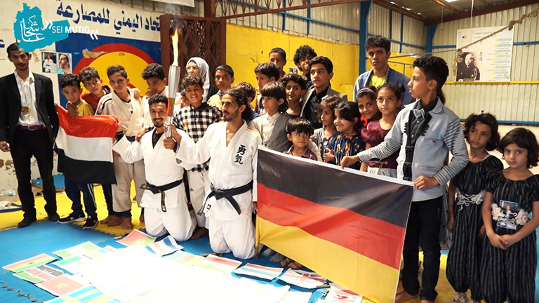 Shugga Nashwan und andere haben sich für ein Gruppenfoto während des Judo-Events in Sanaa aufgestellt. Er kniet, hinter ihm stehen viele Kinder. Die jemenitische und deutsche Flagge sind zu sehen.