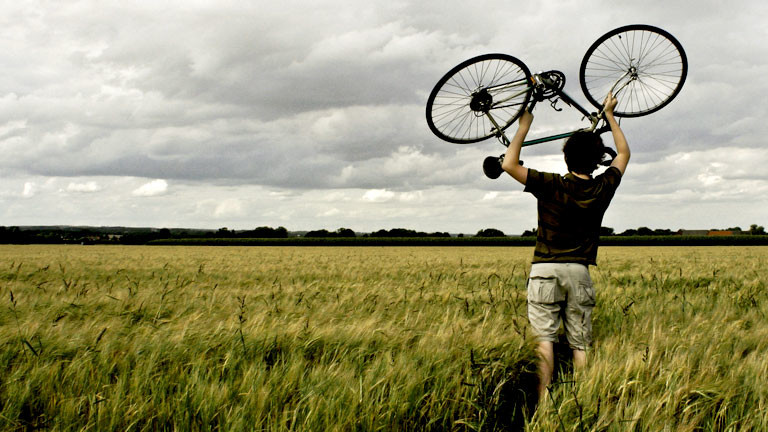 Ein Mann steht in einem Feld und hält ein Fahrrad über den Kopf.