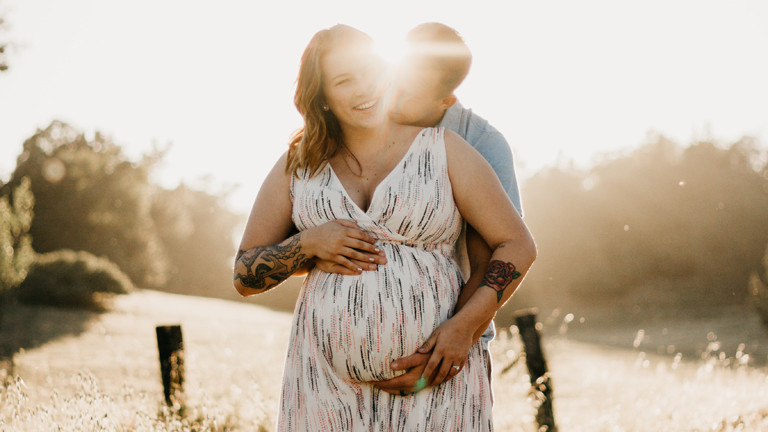 Paar: vorne die schwangere Frau, hinten der Mann, der ihr um den Bauch fasst.