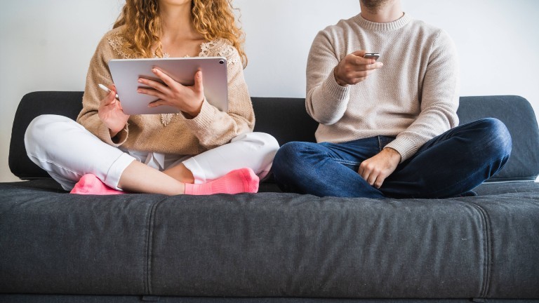 Mann und Frau sitzen im Schneidersitz auf einem Sofa. Die Frau hält ein Tablet in den Händen, der Mann eine Fernbedienung.